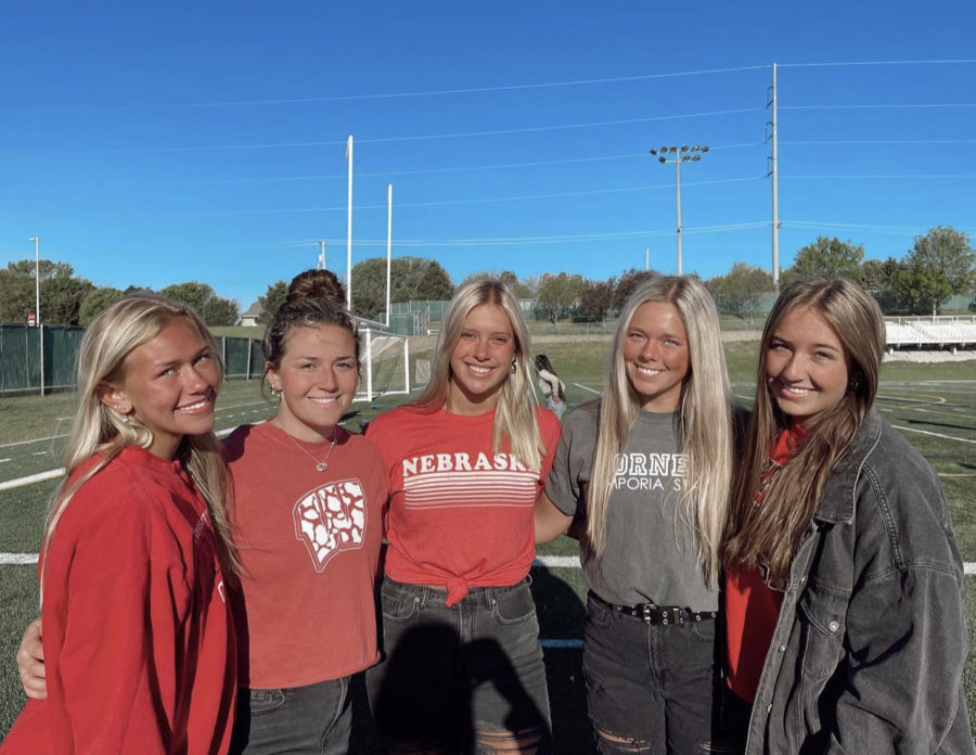 Seniors Dana Summers, Olivia Petersen, Carli Houfek, Jaya VanAckeren and Megan Forst pose together on the turf field with their college shirts representing where they will attend. I will forever be grateful for my time at Millard West, Houfek said. I am excited to start a new chapter, but will definitely miss my friends.