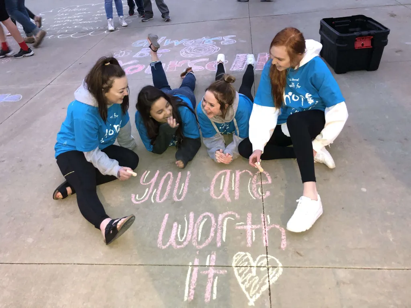 Teenagers from the Out of the Darkness walk are writing inspirational quotes on the sidewalk and talking. 
