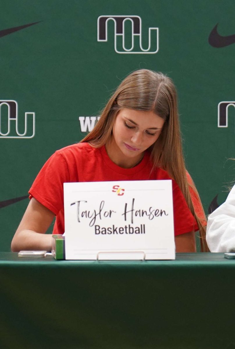 Looking down at a paper, senior Taylor Hansen signs her letter of intent. Hansen was signing to play division three womens basketball. I chose Simpson College in Iowa, Hansen said. Their program really stood out to me because the coaches and players invited me in and welcomed me like I was a part of their family.